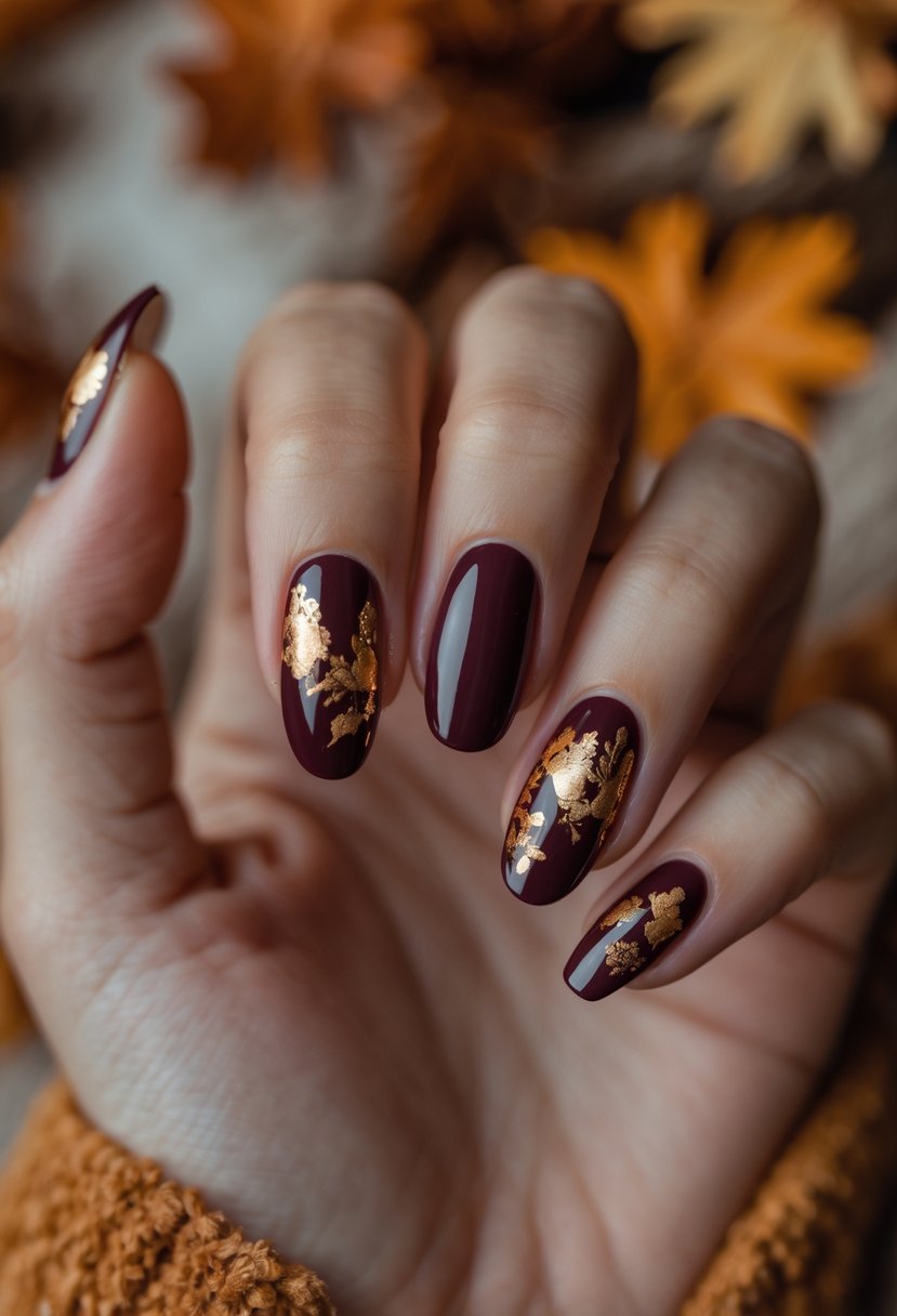 A close-up of a hand with deep burgundy nails decorated with gold foil accents against a warm autumn-colored background.